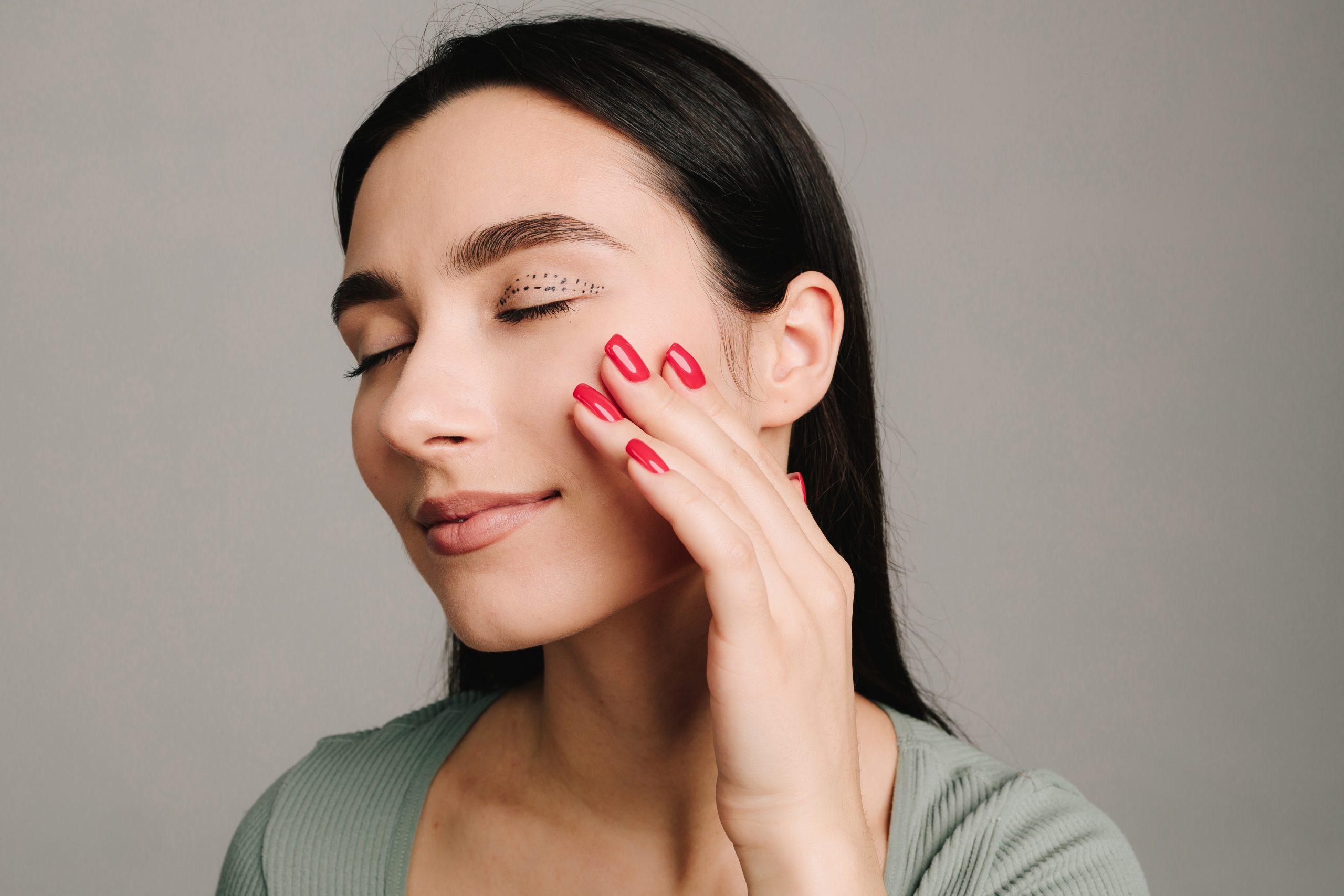 Woman with eyes closed touching her cheek with red nail polish