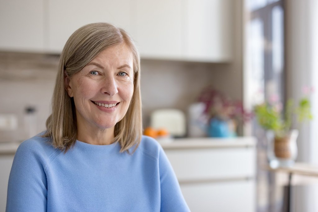 Smiling woman with shoulder-length blonde-gray hair in a blue sweater in a bright kitchen