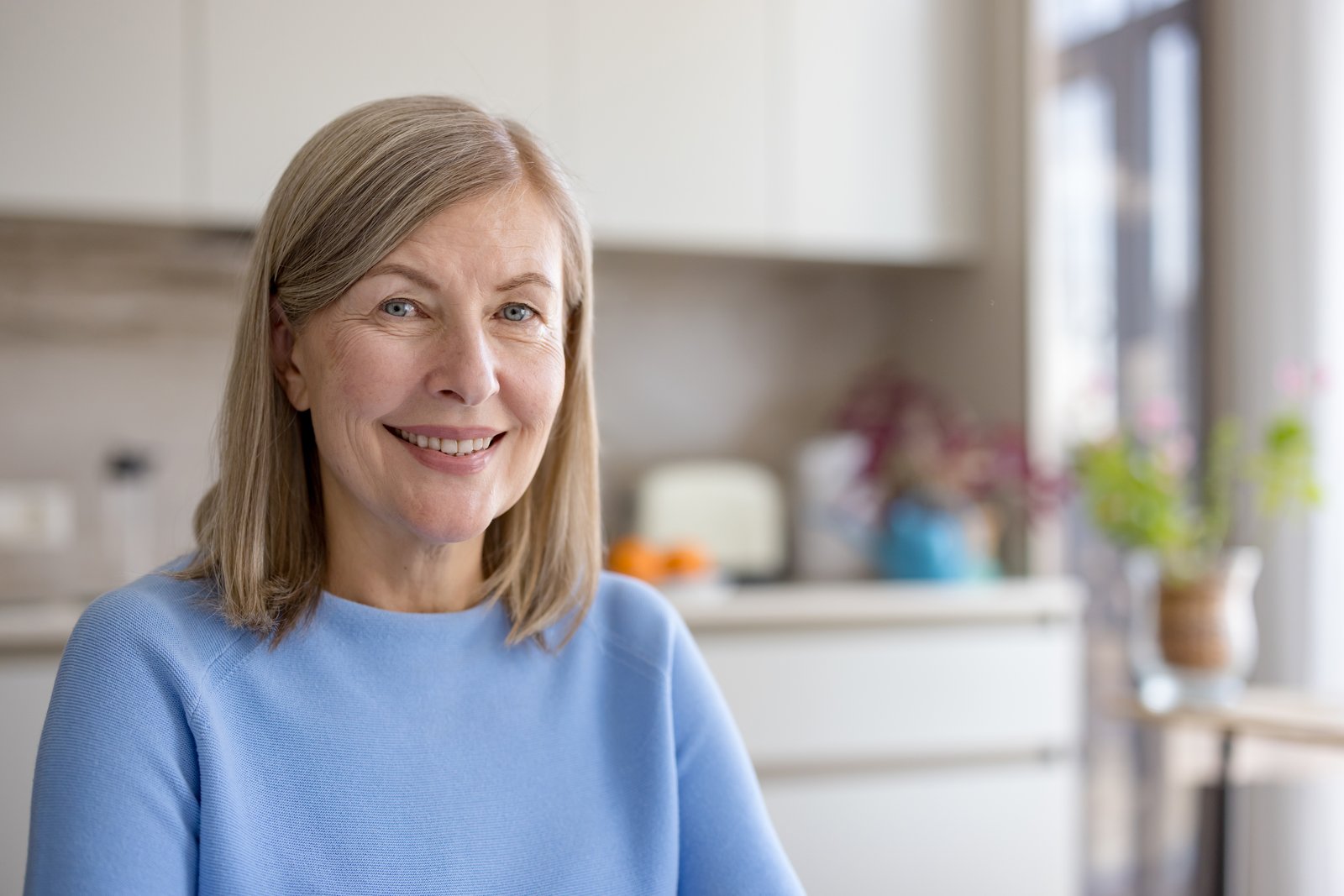Smiling woman with shoulder-length blonde-gray hair in a blue sweater in a bright kitchen