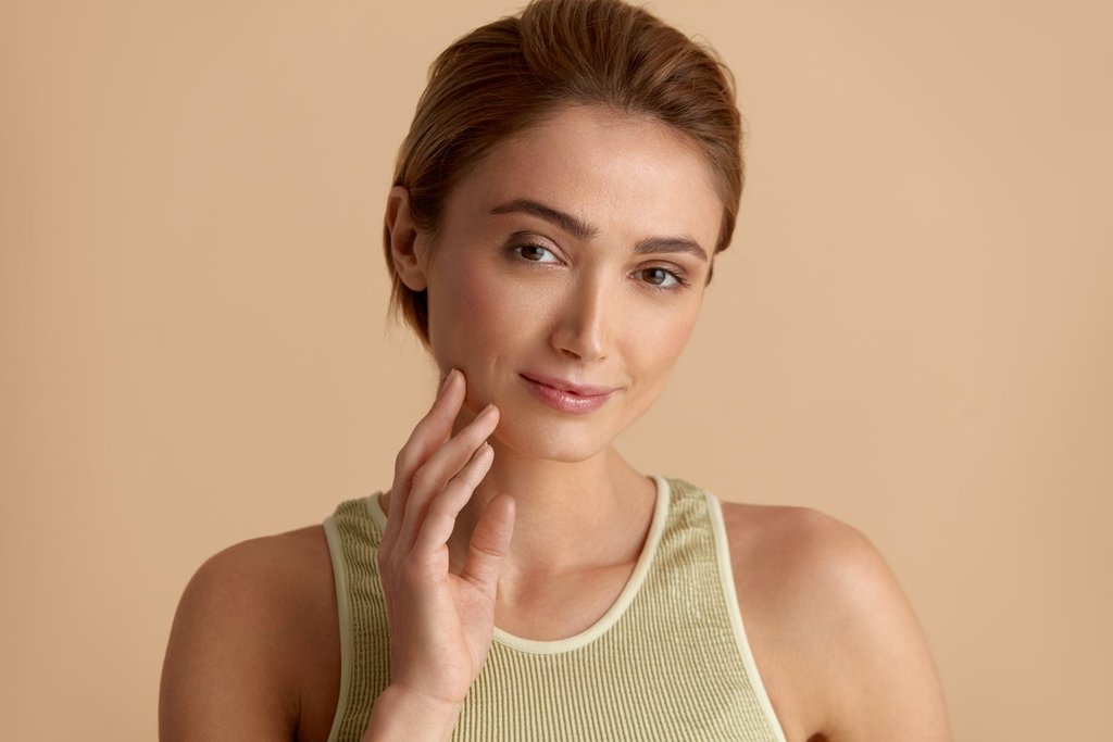 Woman with light-brown hair pulled back, in a sage-green tank top, resting her fingertips on her jawline against a beige background