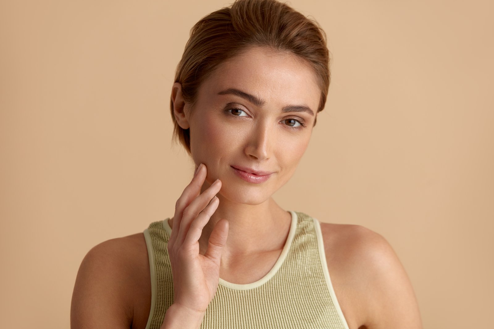 Woman with light-brown hair pulled back, in a sage-green tank top, resting her fingertips on her jawline against a beige background