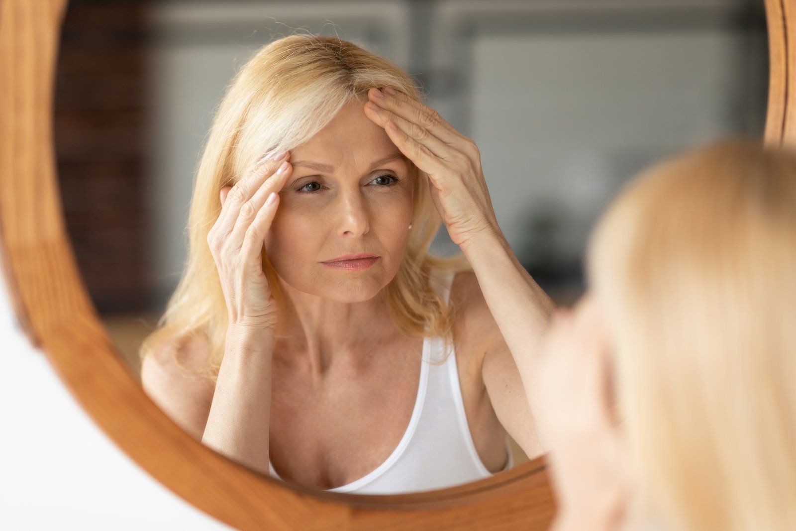 Woman with shoulder-length blonde hair in a white tank top pressing her fingertips to her forehead while looking into a round wood-framed mirror