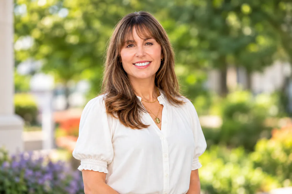 Smiling woman with long brown hair in a white blouse standing outdoors in a sunlit garden
