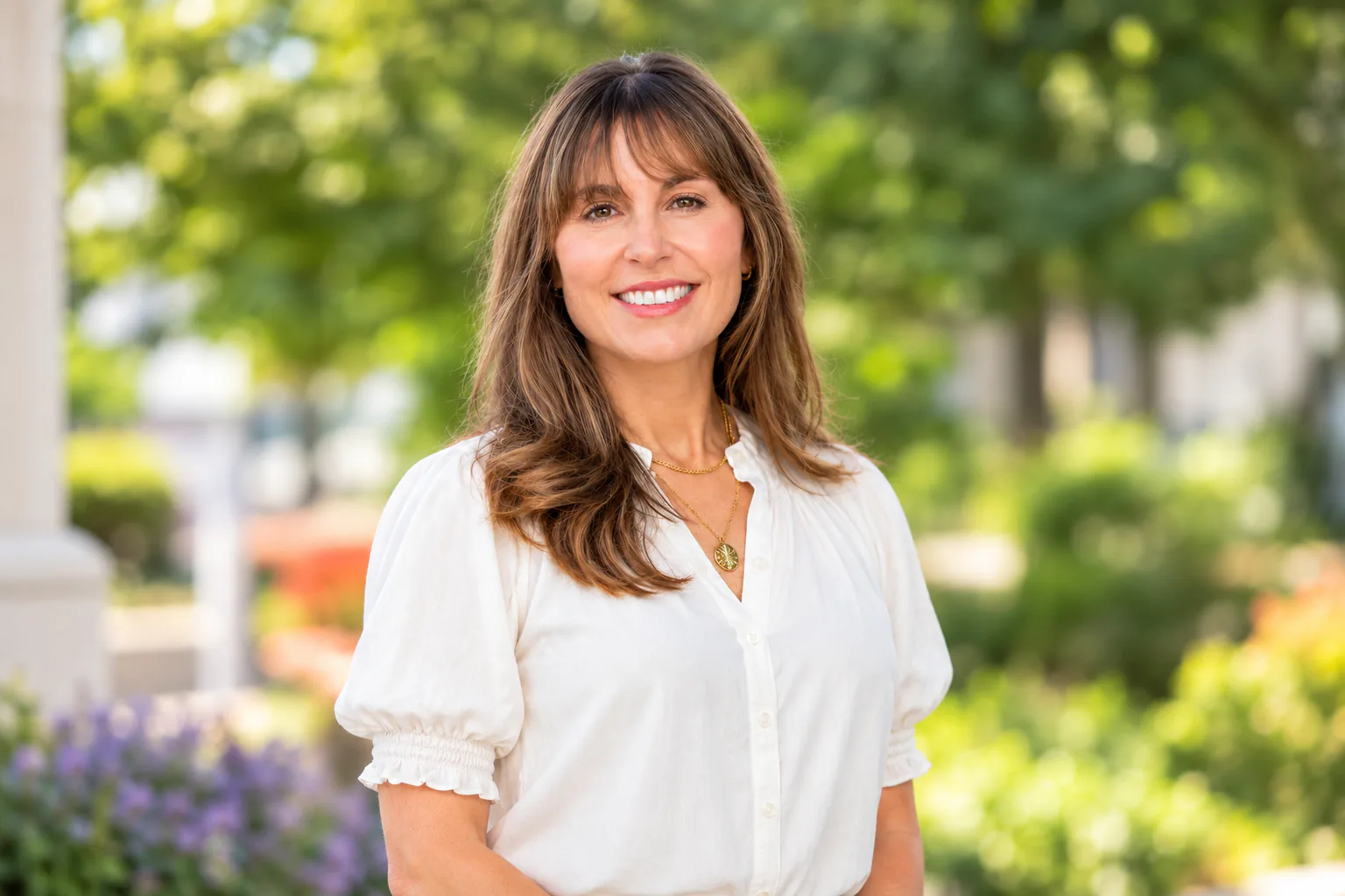 Smiling woman with long brown hair in a white blouse standing outdoors in a sunlit garden