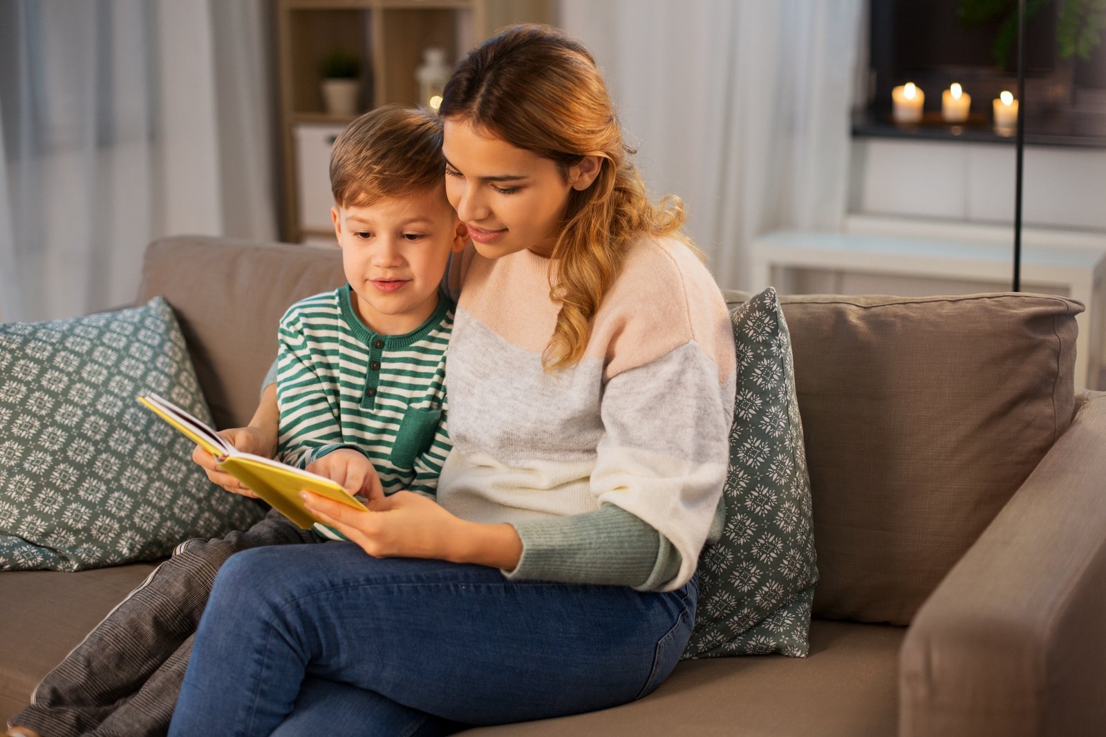 Young woman in a pink and grey sweater reading a yellow book with a small boy in a green striped shirt on a sofa at home