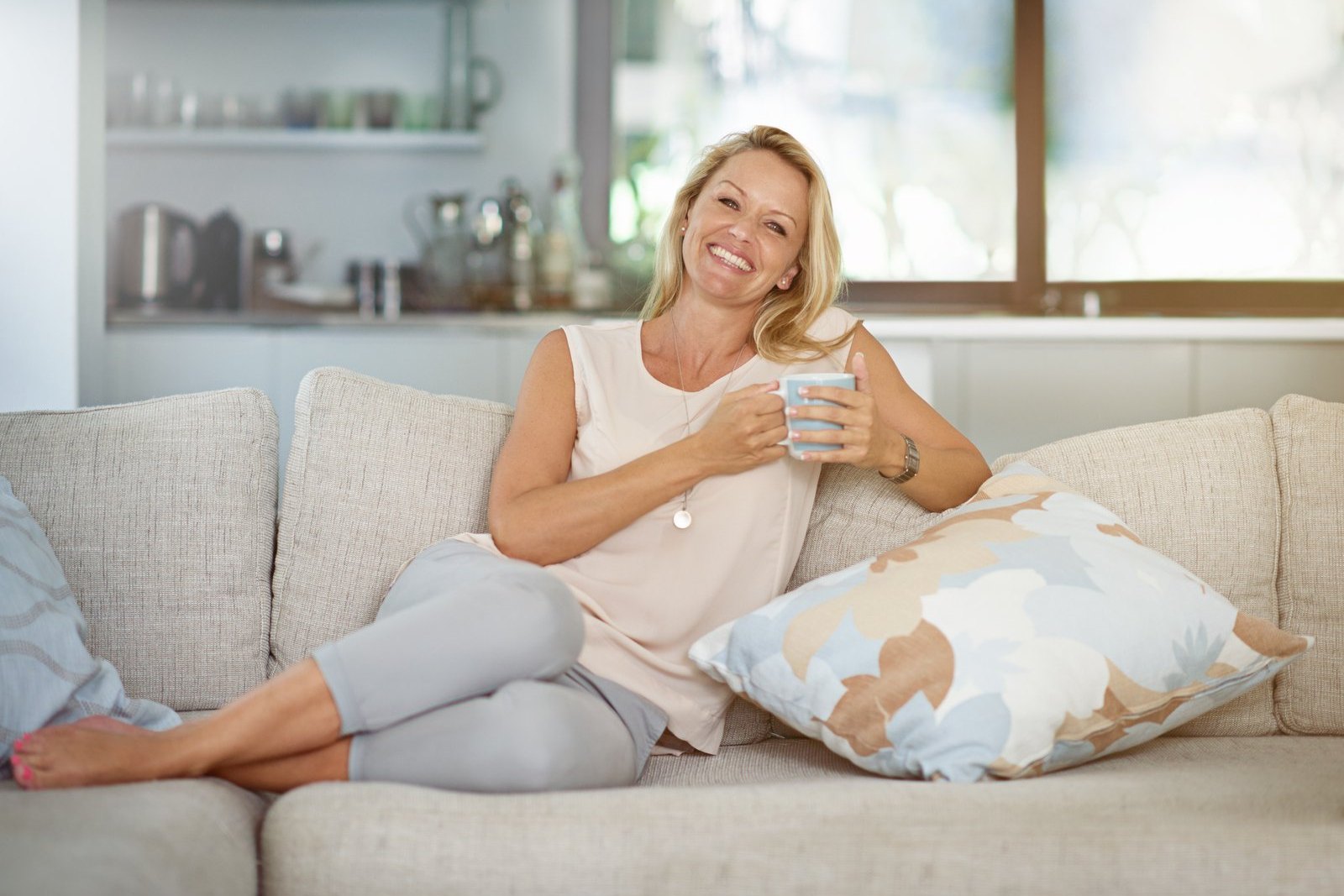 Smiling woman with shoulder-length blonde hair sitting barefoot on a beige sofa, holding a light blue mug beside a floral cushion